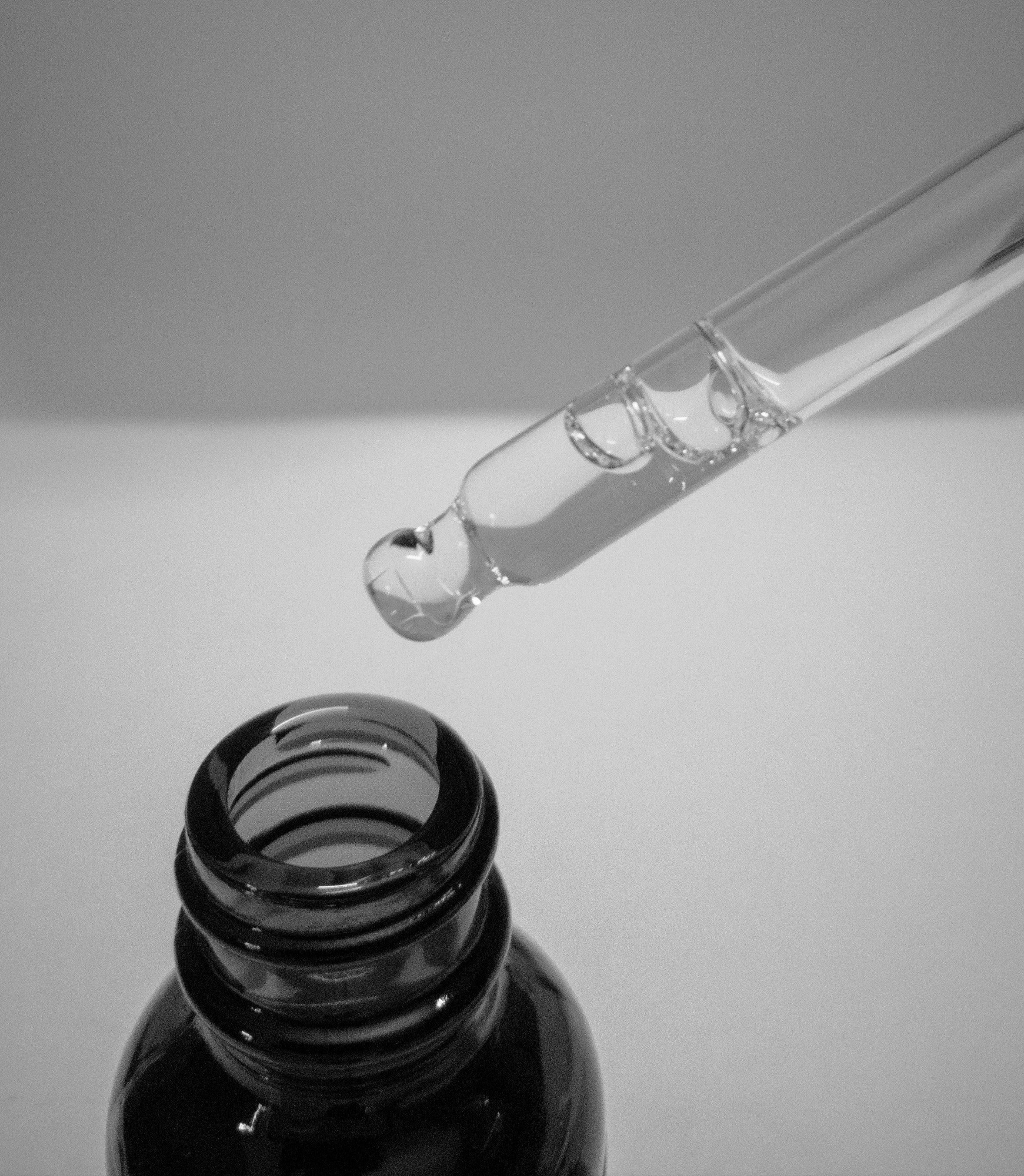 Glass dropper with a drop of liquid over a black bottle on a gray background
