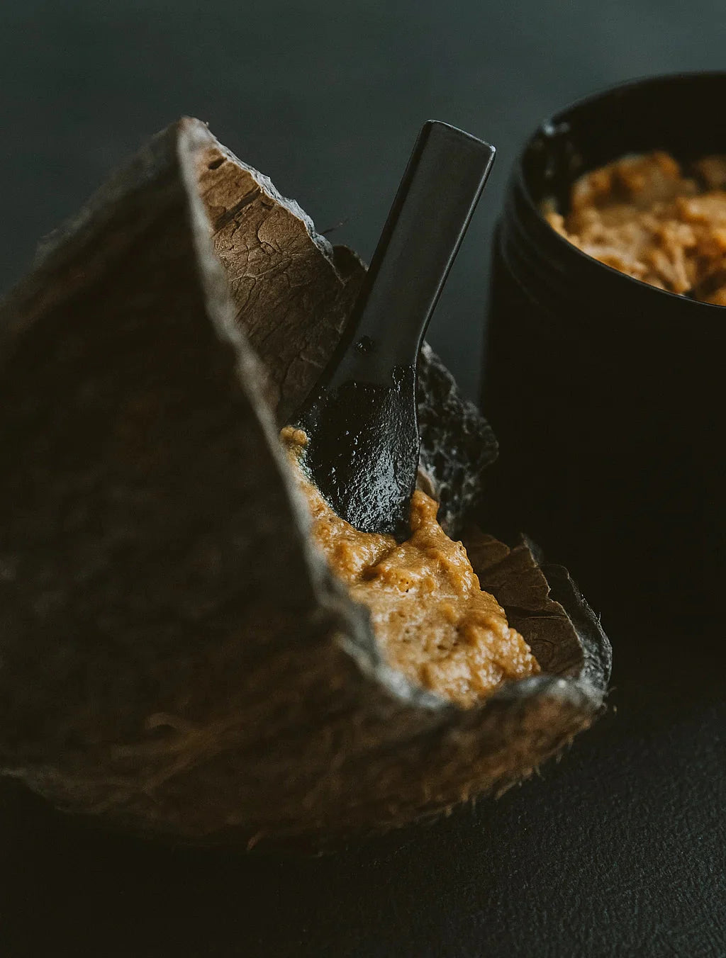 Open coconut filled with brown paste with a spoon inside on a dark background
